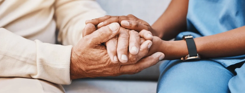 Two people hold hands in a comforting gesture: an older adult's hands resting on a younger caregiver's hands wearing a blue uniform.—care and support scene